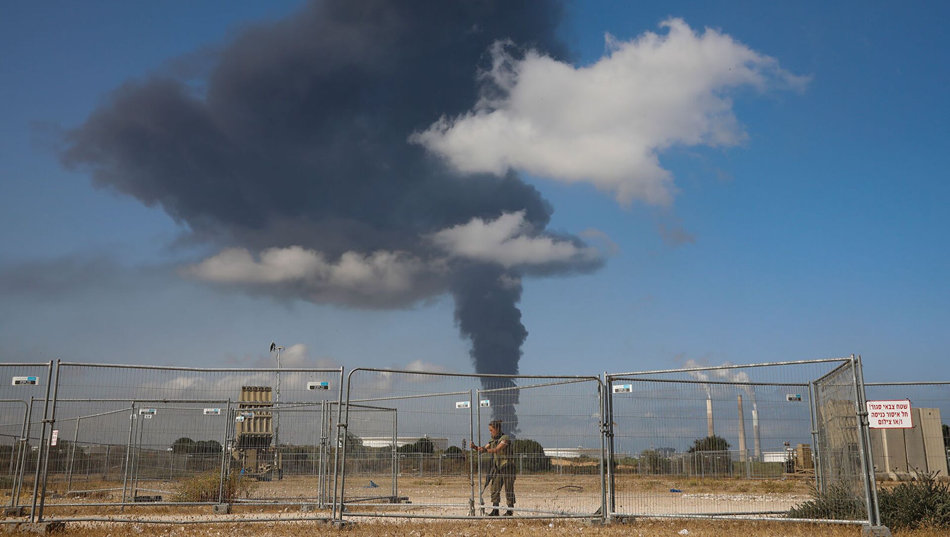 Израильский солдат рядом с системой ПВО Железный купол на фоне горящего нефтяного резервуара, пораженного ракетным обстрелом из сектора Газа (12 мая 2021). Ашкелон - Sputnik Армения, 1920, 12.05.2021
