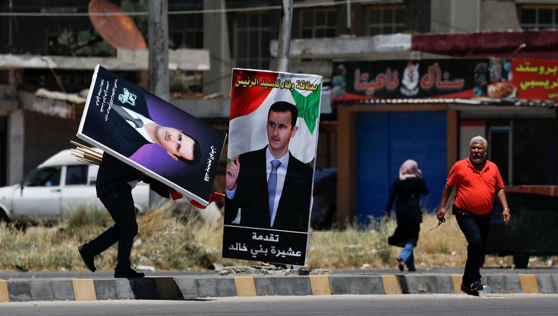 Мужчина идет с плакатами с изображением президента Сирии Башара аль-Асада в преддверии президентских выборов 26 мая в районе Аль-Ваер в Хомсе - Sputnik Армения, 1920, 10.08.2021