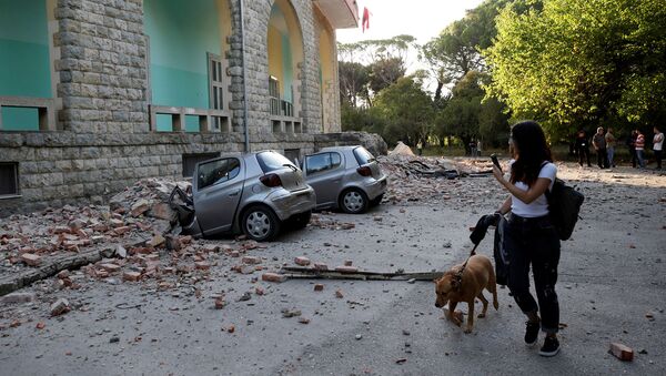 Женщина с собакой проходит мимо разрушенных автомобилей и поврежденного здания после землетрясения (21 сентября 2019). Тиранa - Sputnik Արմենիա