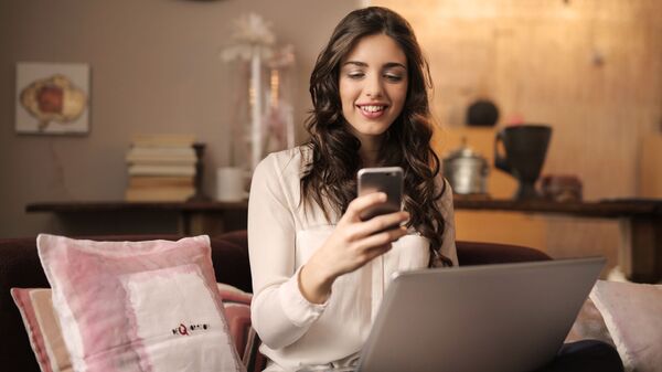 Симпатичная девушка брюнетка сидит дома за ноутбуком и улыбаясь смотрит в свой смартфон - Sputnik Армения