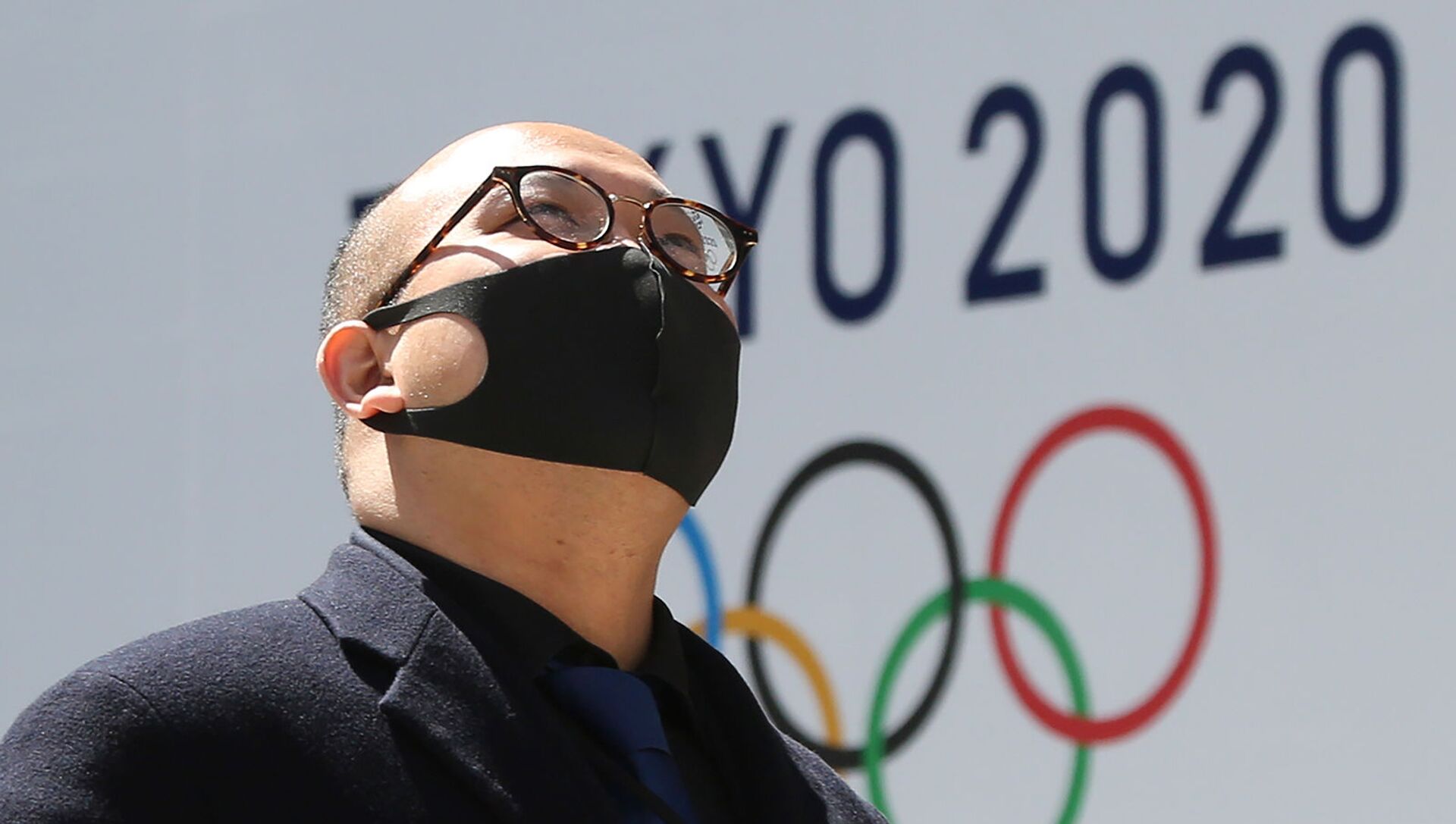 Мужчина в защитной маске проходит перед логотипом токийской Олимпиады (25 марта 2020). Токио - Sputnik Армения, 1920, 31.07.2021